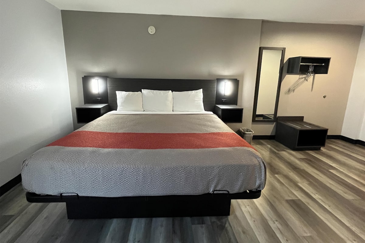 Ein Hotelzimmer mit einem großen Bett, zwei Nachttischen mit Lampen, einer kleinen Mülleimer und einem Spiegel an der Wand.