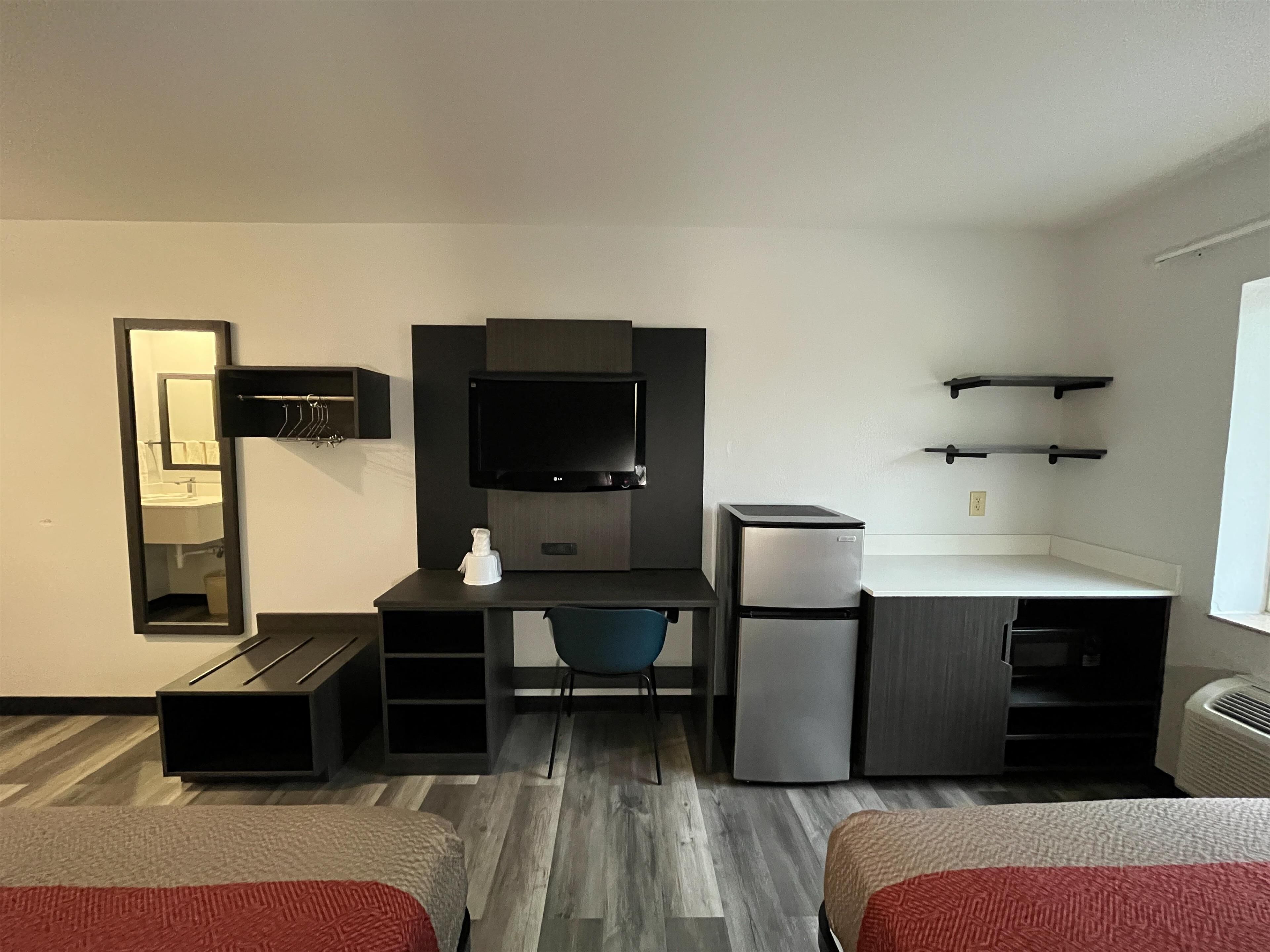 Una habitación de hotel moderna con escritorio, silla y un refrigerador pequeño. Cuenta con una cama con manta roja, un espejo y un estante con artículos de aseo.