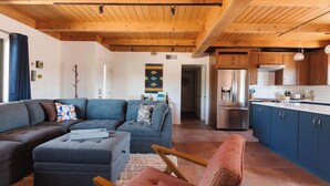 A modern living room with a grey sectional sofa, a wooden coffee table, and a red armchair. A kitchen with blue cabinets and stainless steel appliances is visible in the background.