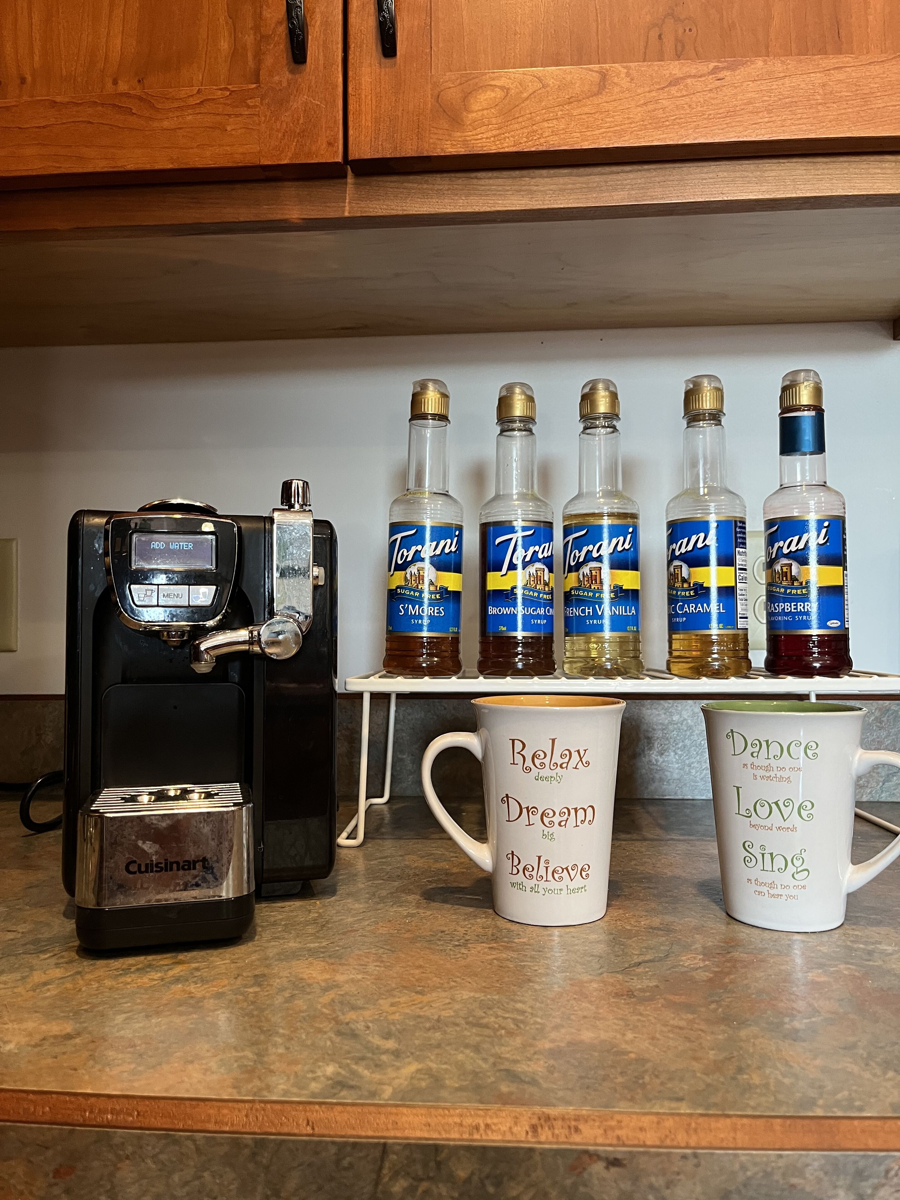 A kitchen counter with a coffee machine, sugar-free syrup bottles, and two mugs with inspirational phrases.