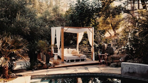 A secluded outdoor bedroom with a canopy bed, surrounded by lush vegetation and a swimming pool.