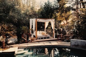 A secluded outdoor bedroom with a canopy bed, surrounded by lush vegetation and a swimming pool.
