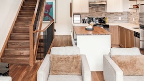 A modern kitchen with a central island, wooden staircase, and a view of the living area with two sofas. - East End Loft (Calgary)