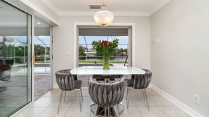 A dining area with a round table, four upholstered chairs, a chandelier, and a view of a pool and greenery outside. - La Dolce Vita: Your Waterfront Jackpot (North Fort Myers)