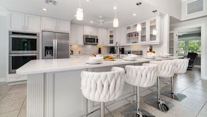 A modern kitchen with white cabinetry, stainless steel appliances, a large island with bar stools, pendant lighting, and a view to the outside. - La Dolce Vita: Your Waterfront Jackpot (North Fort Myers)