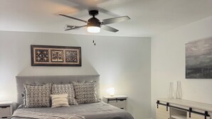 Une chambre à coucher comprenant un lit, un ventilateur de plafond, une table de chevet, une commode et un tableau encadré accroché au mur.