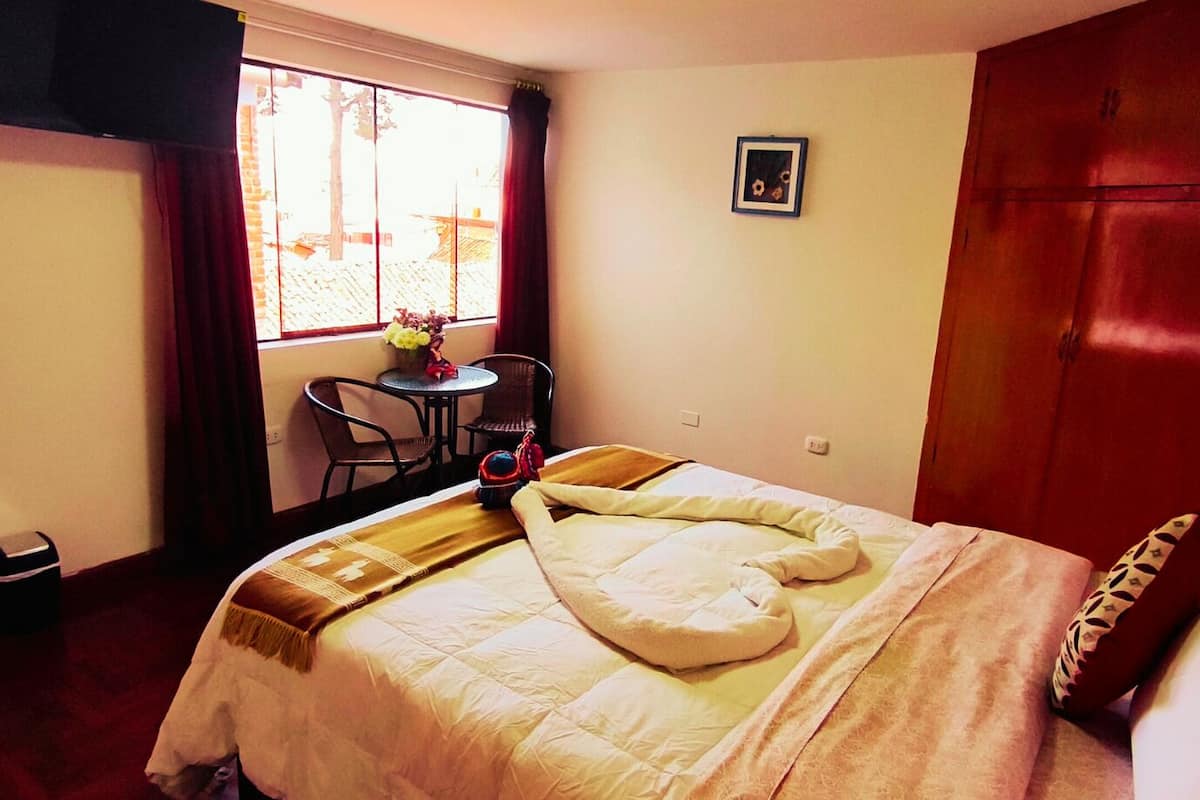 A hotel room with a bed, a small table with chairs, a window with curtains, and a door.