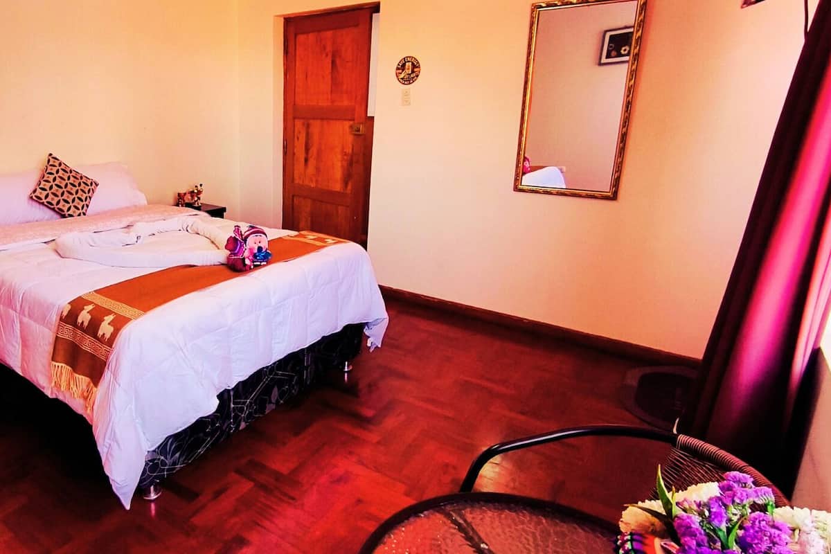 A bedroom with a bed, a wooden door, a mirror, and a chair with a flower arrangement.