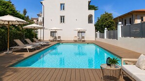 A swimming pool with lounge chairs and umbrellas, a white building with a balcony, and a fenced area with a sign. - Primo Hotel (Marciana Marina)