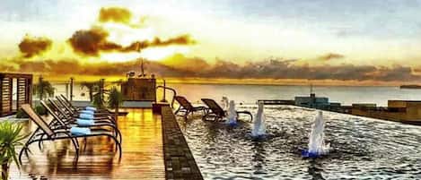 A pool area with lounge chairs and a view of the sea during sunset.