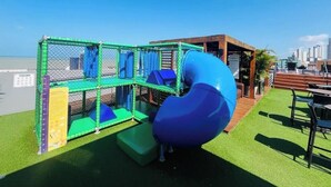 A playground with a slide and netted play area, located on a grassy deck with a view of the city skyline. - LM Quality Suites (João Pessoa)