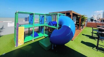 Un parque infantil con tobogán y zona de juegos con red, situado en una terraza ajardinada con vistas al horizonte de la ciudad.