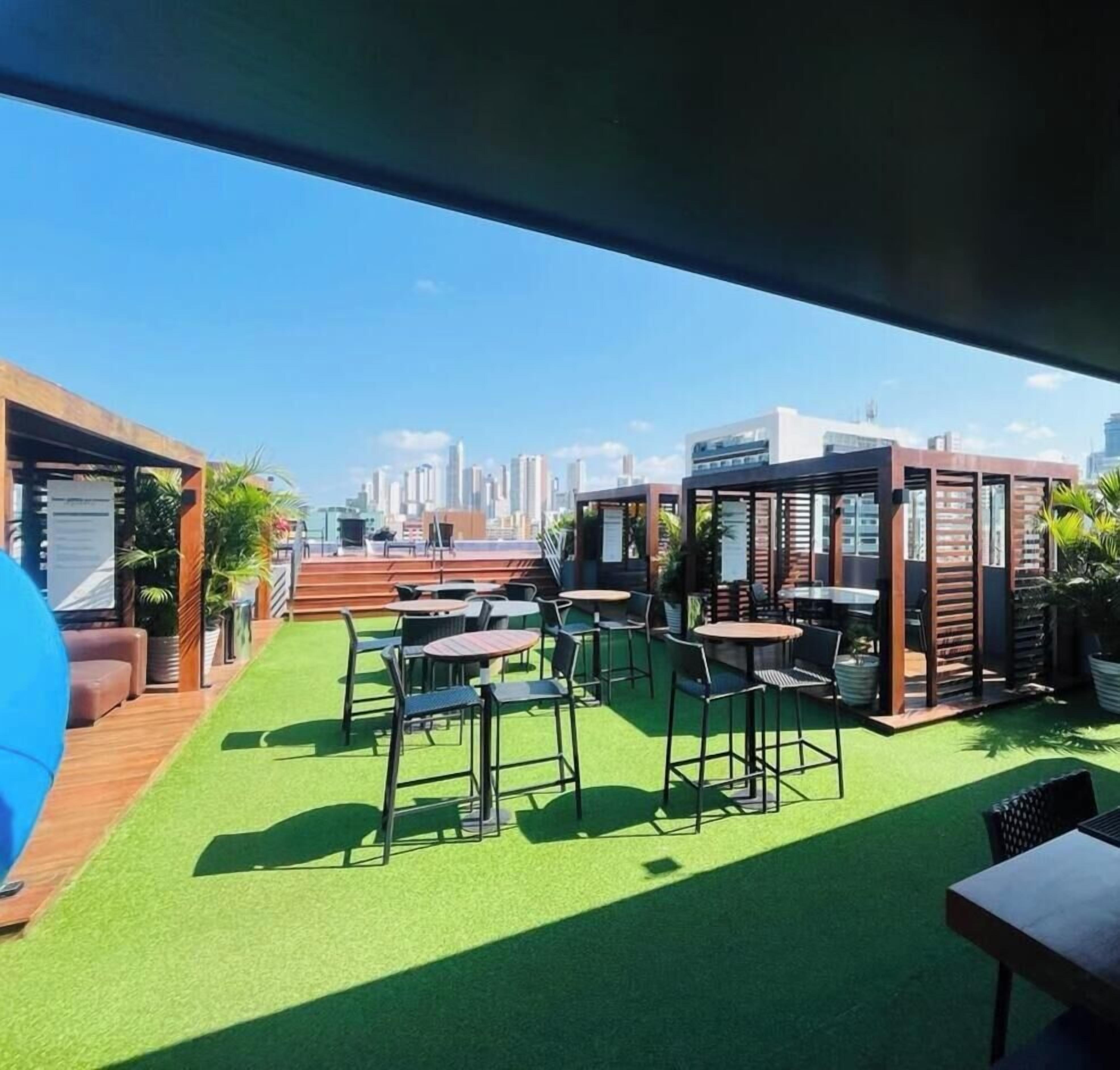 An outdoor seating area with tables and chairs, a green artificial turf, and a city skyline in the background.