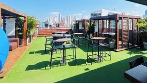 An outdoor seating area with tables and chairs, a green artificial turf, and a city skyline in the background. - LM Quality Suites (João Pessoa)