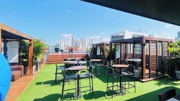 An outdoor seating area with tables and chairs, a green artificial turf, and a city skyline in the background.
