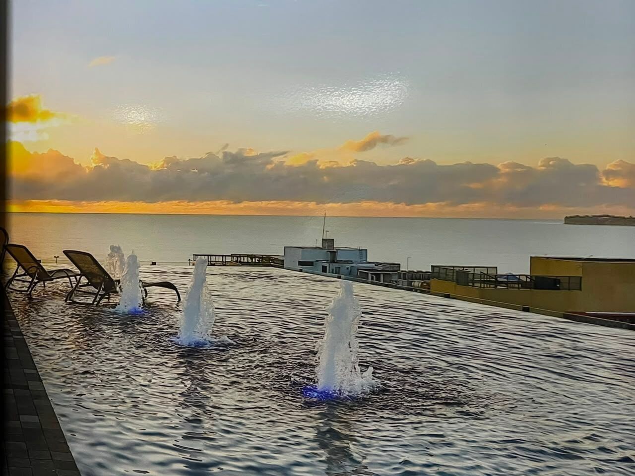 A pool with water jets, lounge chairs, and a view of the sea at sunset.