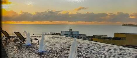 Una piscina con chorros de agua, sillas de descanso y vistas al mar al atardecer.