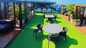 Un espace extérieur aménagé en terrasse, avec une pelouse artificielle verte, des tables et des chaises rondes, un coin bar et une aire de jeux pour enfants.