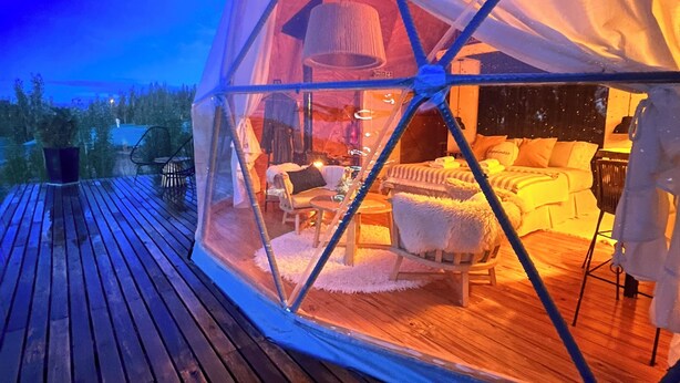A dome-shaped tent with a wooden deck outside, furnished with a bed, chairs, and a table.