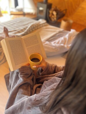 Una persona leyendo un libro mientras sostiene una taza, sentada en una cama con una manta.