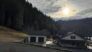 A two-story house with a garage, surrounded by a forested hillside. - Selway Chalet (Kooskia)