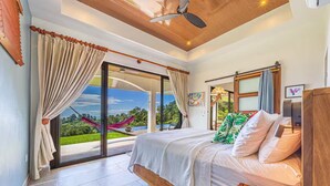 Une chambre à coucher avec un lit, un ventilateur de plafond et une vue sur un jardin et le ciel, grâce à de grandes fenêtres.