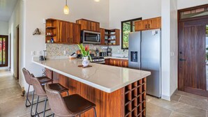 Une cuisine moderne avec des meubles en bois, un grand îlot central, des appareils électroménagers en acier inoxydable et une vue sur l’extérieur.