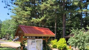 A wooden cabin with a red roof, surrounded by rocks and plants, with a 'SLOW' sign nearby. - Jack Pine Cabin at Pine Ridge Park (Pembroke)