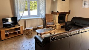 A living room with a fireplace, a television, a sofa, and a window with curtains. - Family house with terrace in Fréhel | 8 guests | Ref 301 (FREHEL)