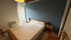 A bedroom with a bed, bedside tables, a chair, a window, and a hanging light fixture. - Family house with terrace in Fréhel | 8 guests | Ref 301 (FREHEL)