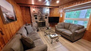Indbydende interiør i en hytte med en pejs af natursten, to grå sofaer, et sofabord og et stort vindue med udsigt over skoven.