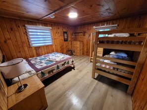 Une chambre dans une cabane en bois, avec un lit superposé, une courtepointe, une commode en bois et une fenêtre avec des stores.