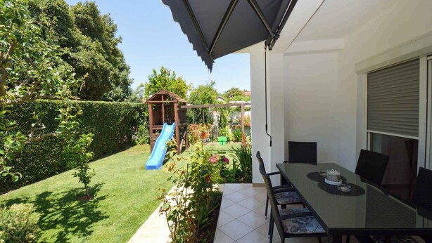 Eine Terrasse mit Tisch und Stühlen, eine Rutsche und ein Garten mit verschiedenen Pflanzen und einer hölzernen Spielanlage.