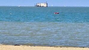 A beach with calm waters, a distant building, and a small red object floating.