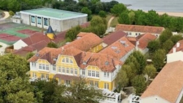 An aerial view of a coastal residential area with houses, a sports field, and a beach in the background.