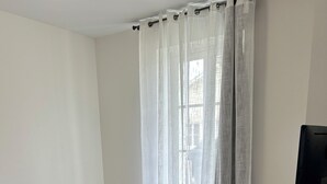 A bedroom with a bed, a television on a stand, and a window with sheer curtains.