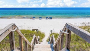 A wooden boardwalk leading to a beach with clear blue water and a few umbrellas.