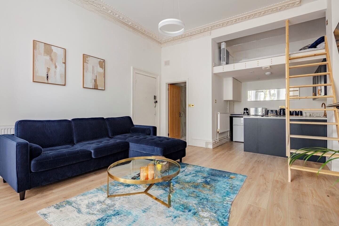 A modern living room with a dark blue sofa, a glass coffee table, and a blue patterned rug. A kitchen with white cabinets and a wooden ladder is visible in the background.