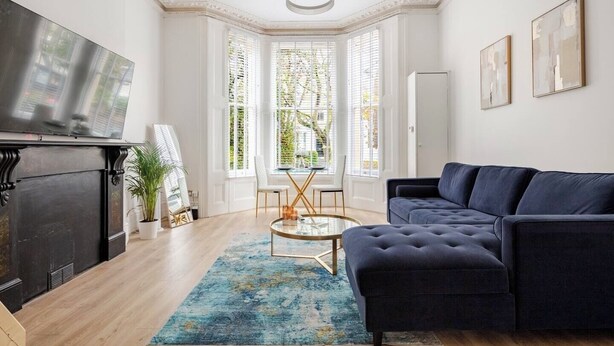 Una sala de estar moderna con un sofá azul oscuro, una mesa de centro de vidrio y un tapete azul. Hay una chimenea con un televisor encima y una ventana grande con persianas.