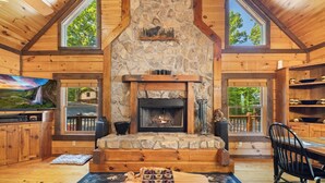 L’intérieur chaleureux d’un chalet avec une cheminée en pierre, des murs en bois et de grandes fenêtres offrant une vue sur le paysage extérieur.
