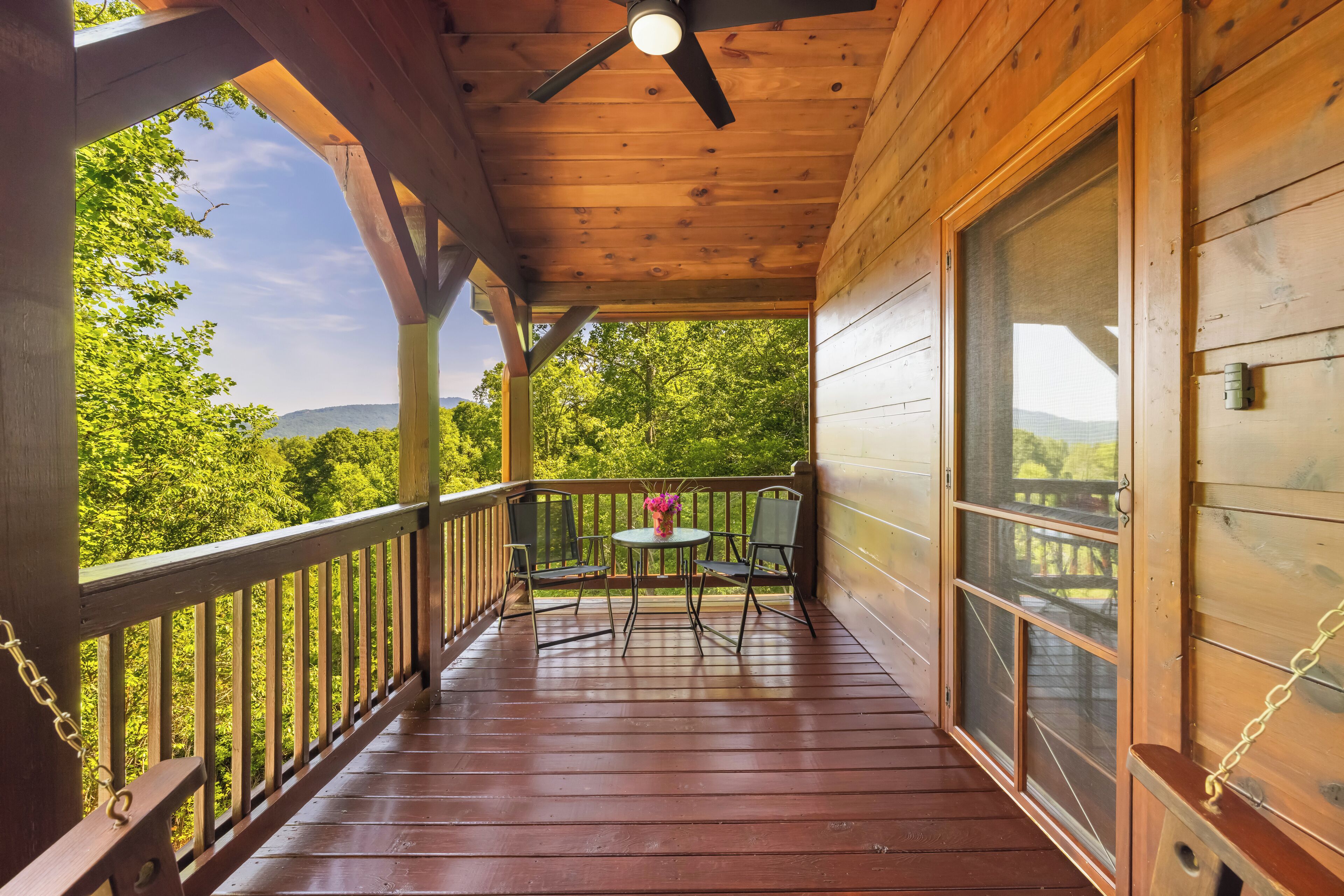 A wooden deck with a ceiling fan, a table with chairs, and a view of green trees and hills.