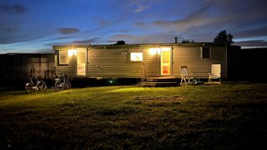 Une maison mobile avec une véranda et deux chaises, un vélo et une pelouse.
