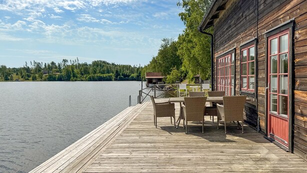 A wooden deck with a table and chairs, overlooking a calm lake with a dock and a building with red-framed windows. - Lovely home in Gränna with house sea view (Gränna)