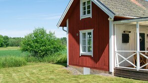 A red wooden house with a white porch and a door, surrounded by a grassy field and trees. - Cozy home in Närkesberg (Närkesberg)