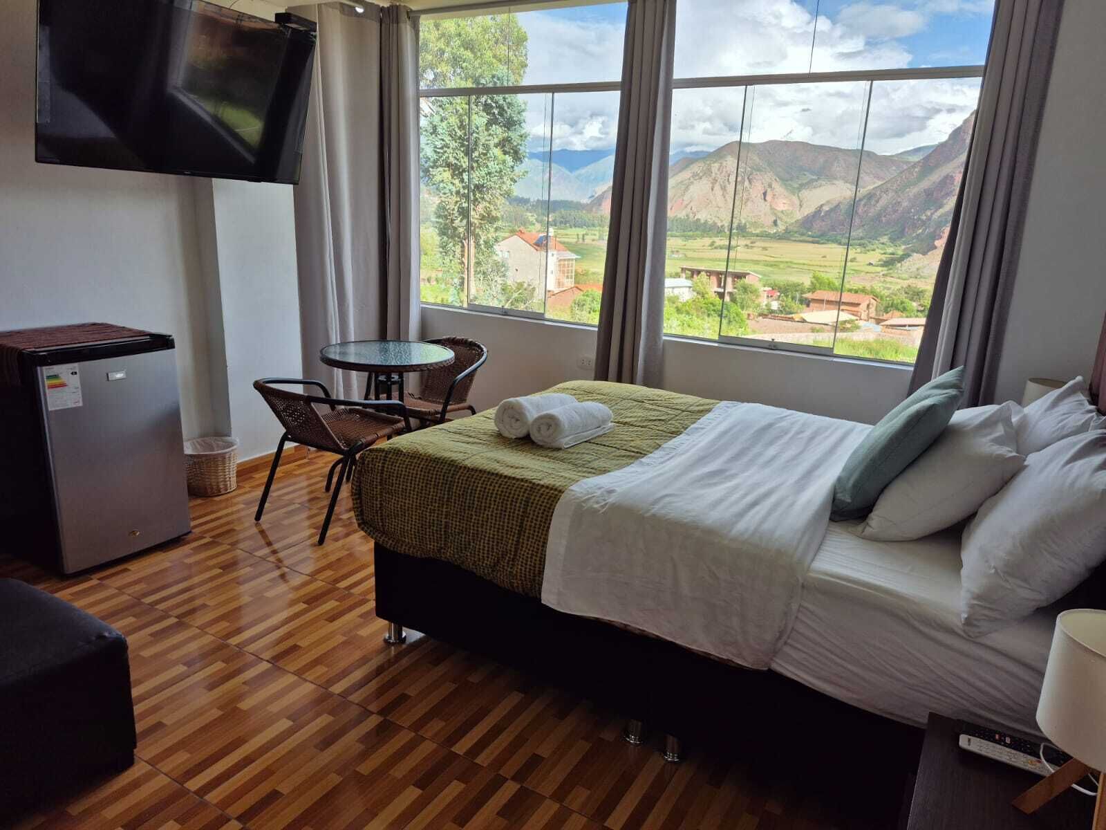 A hotel room with a bed, a small table with chairs, a TV mounted on the wall, and a window with a mountain view.