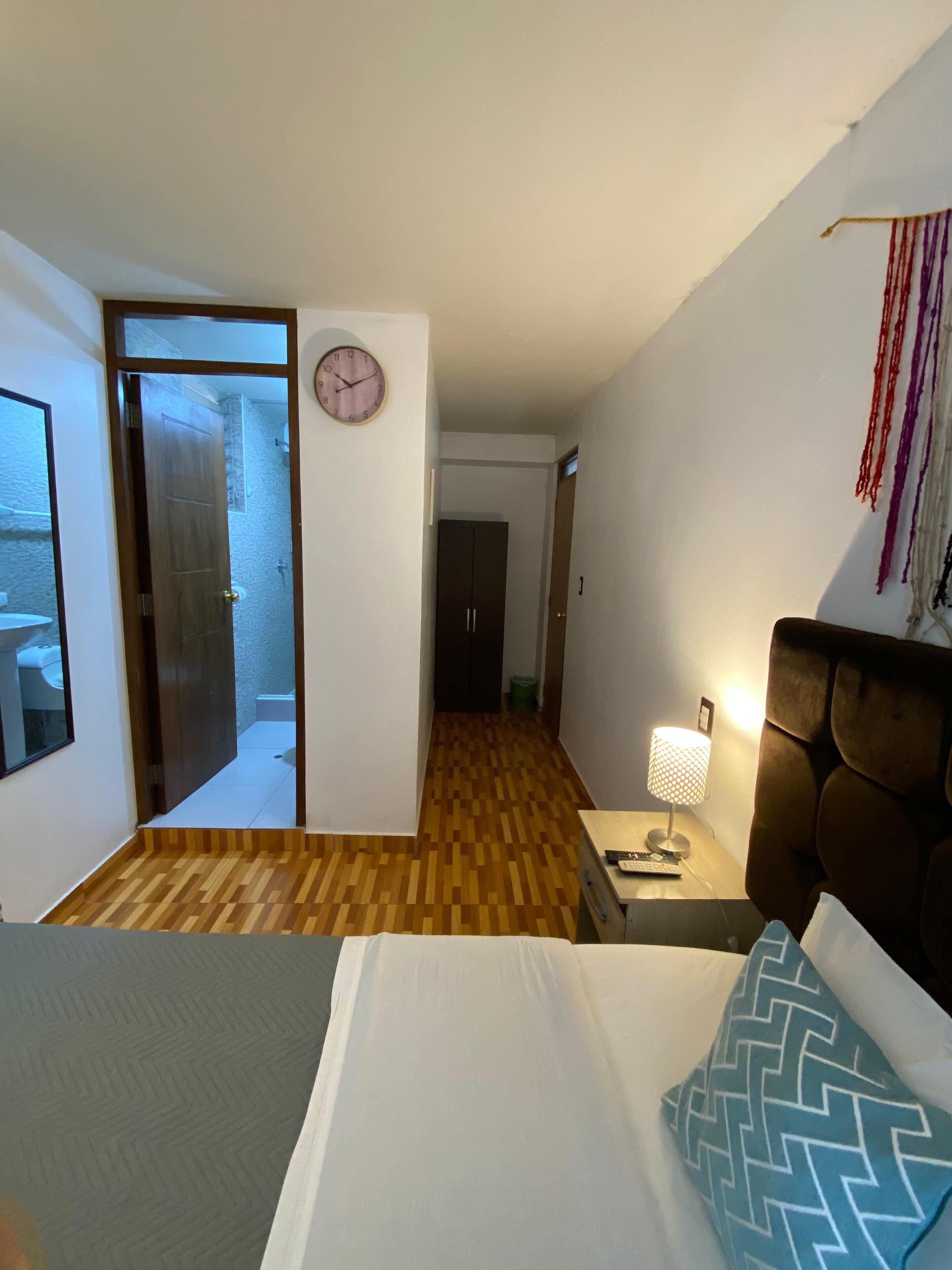 Una habitación de hotel con una cama, una mesita de noche con una lámpara, piso de madera, un reloj de pared y una puerta que da a un baño.