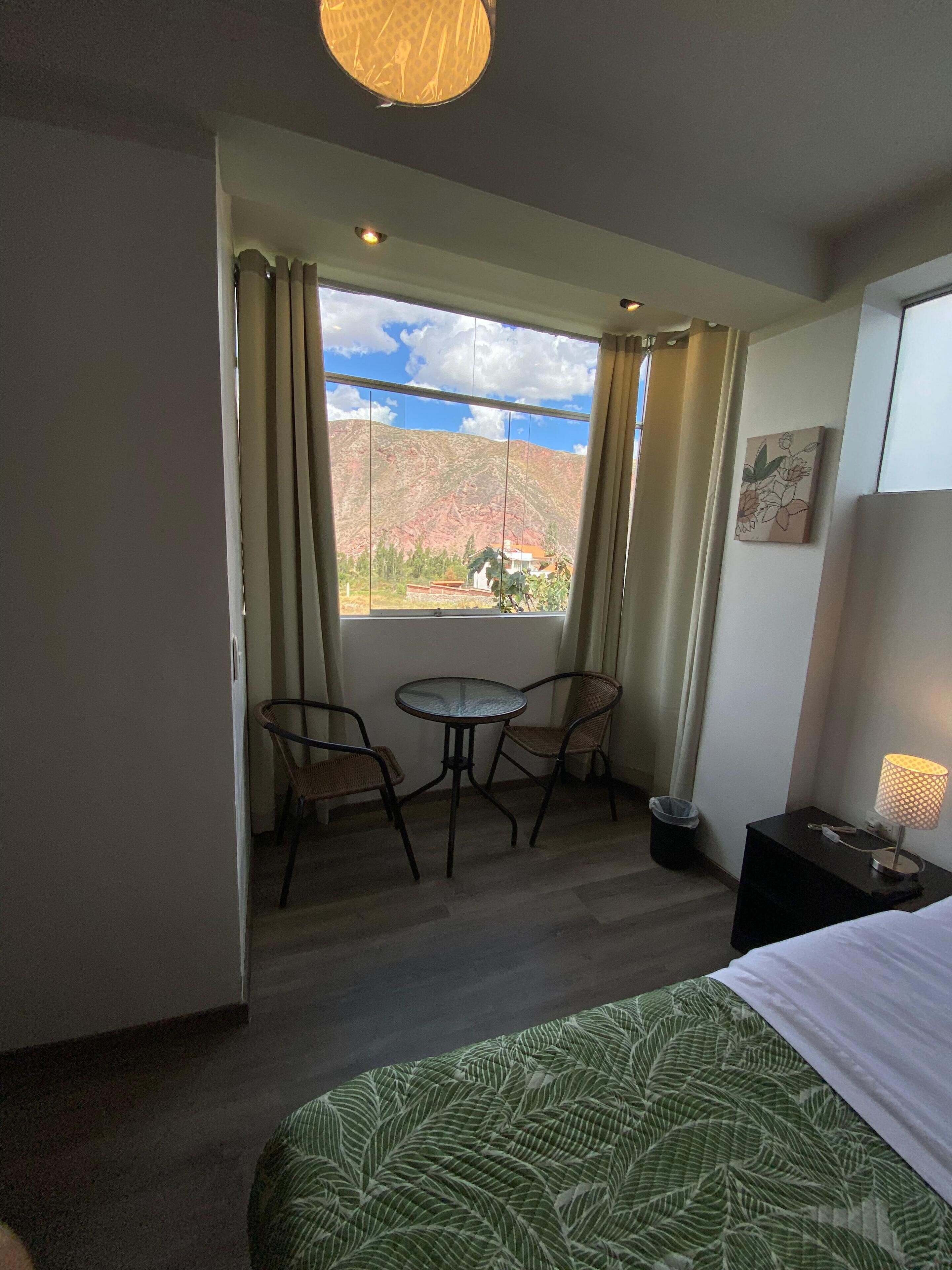 A hotel room with a bed, a small table with two chairs, a window with curtains, a lamp, and a painting on the wall.