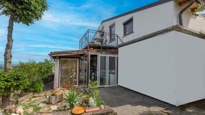 Une maison moderne avec une entrée dotée d’une porte en verre, un petit balcon et un jardin agrémenté de diverses plantes et fleurs.