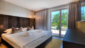 A large bed with multiple pillows, a wooden headboard, and a view of a garden through a sliding glass door.