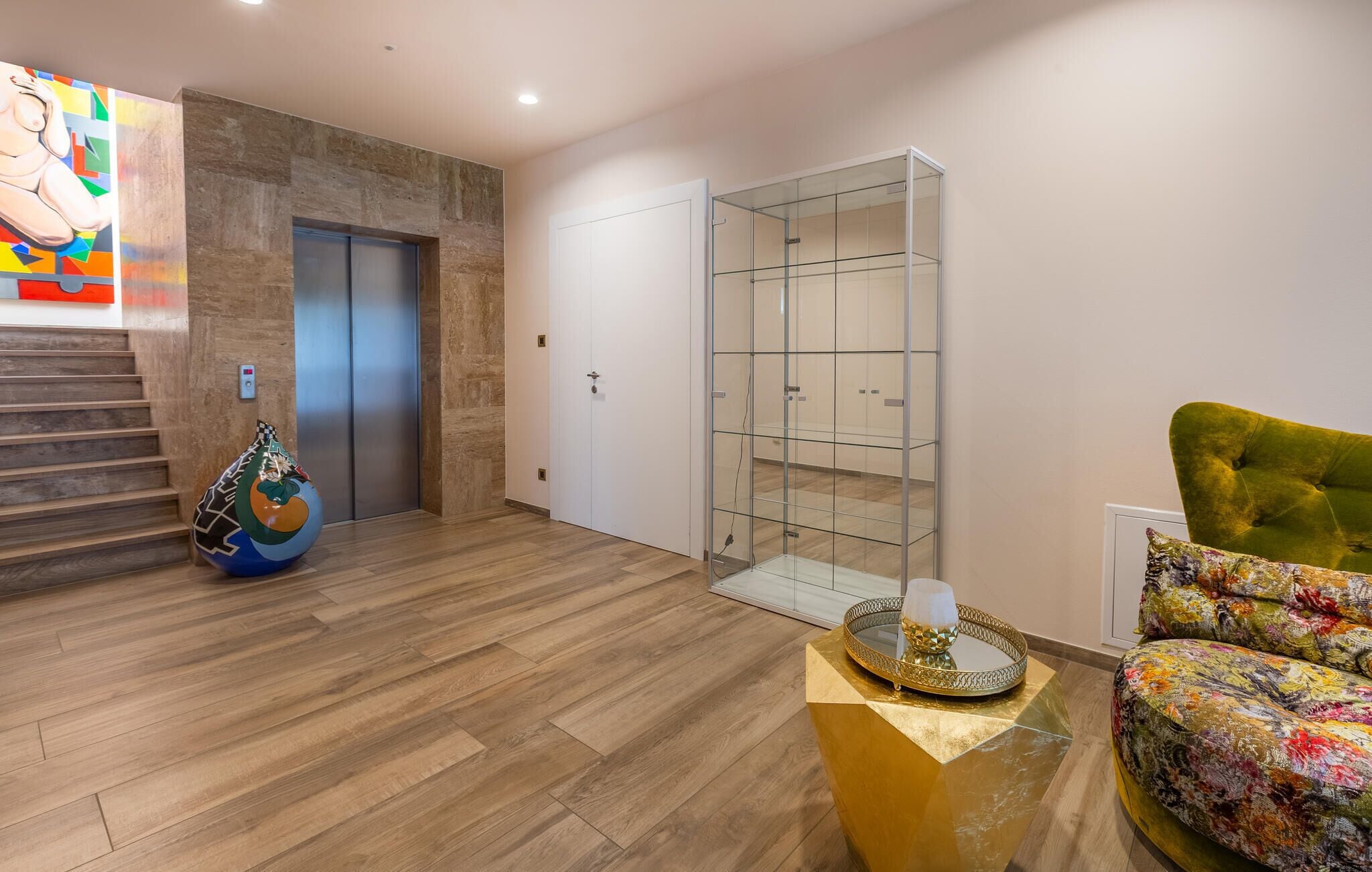 Un salon moderne avec un plancher en bois, un fauteuil recouvert d’un tissu vert, une étagère en verre et un tableau coloré accroché au mur.
